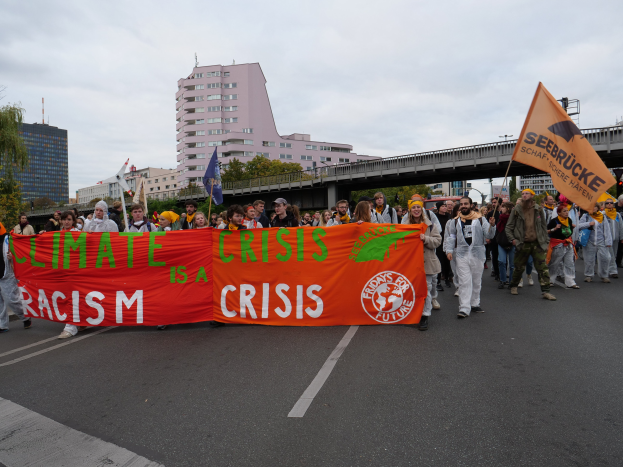 Eine Gruppe von Menschen marschiert eine baumbestandene Straße entlang und hält ein Banner mit der Aufschrift "Klima-Krise ist eine Krise", mit Gebäuden und einer Brücke im Hintergrund unter einem bewölkten Himmel.