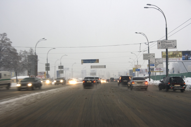 Eine belebte Stadtstraße mit starkem Verkehr an einem verschneiten Tag, mit Fahrzeugen, schneebedeckter Straße, Laternen, Schildern, Bäumen, Gebäuden und einem Himmel im Hintergrund.