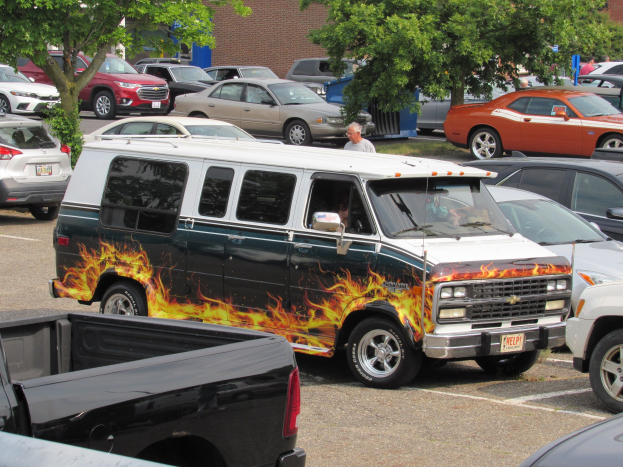 Ein Lieferwagen mit Flammenaufkleber auf der Seite, der auf einem von Bäumen und Gebäuden umgebenen Parkplatz steht, mit Menschen in der Nähe.
