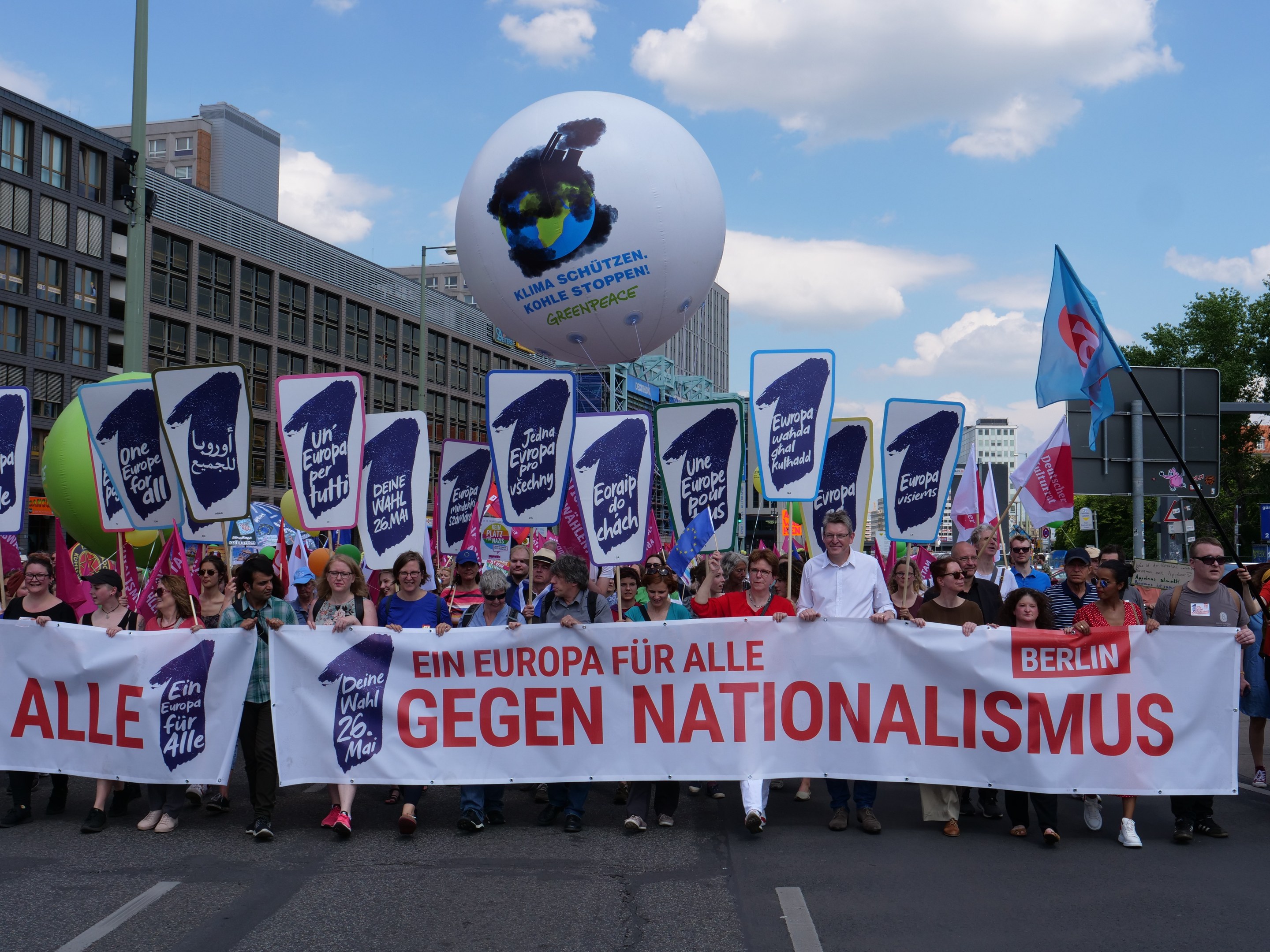 A group of people walking down a street holding a banner that reads "Ein Europa für alle Gegen Nationalismus" and flags, with buildings, trees, and a cloudy sky in the background.