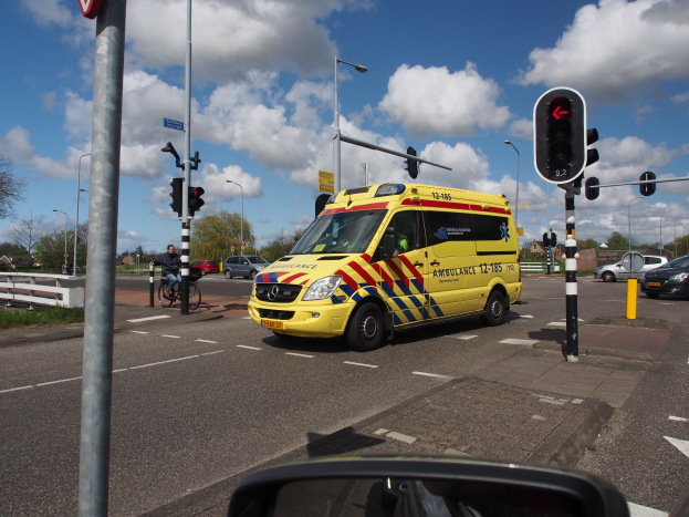 Ambulanz fährt auf einer Straße neben einer Ampel, mit einem Radfahrer im Vordergrund, umgeben von Laternenmästen, Bäumen, Gebäuden und einem bewölktem Himmel, mit einem Spiegel unten.