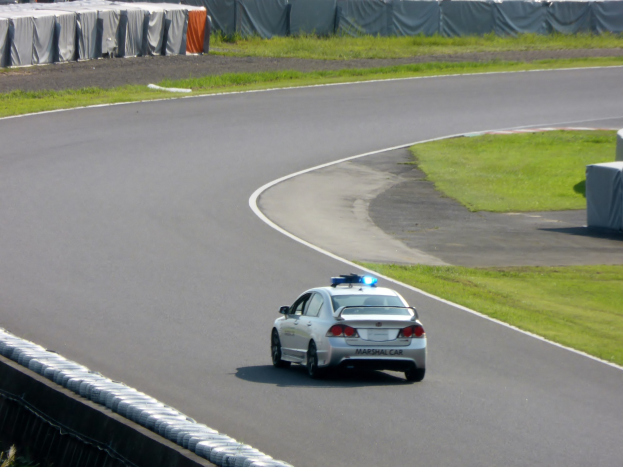Polizeiauto auf einer Rennstrecke mit Gras auf beiden Seiten und Zuschauern und Zelten im Hintergrund.