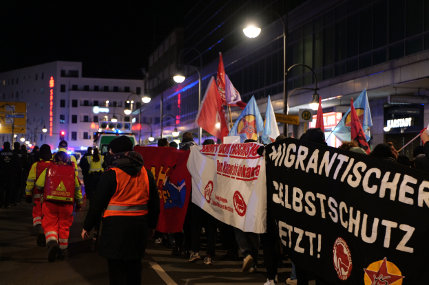 Eine Gruppe von Menschen marschiert nachts in Berlin, trägt Fahnen und Schilder, mit Gebäuden, Straßenlaternen und einem Fahrzeug im Hintergrund.