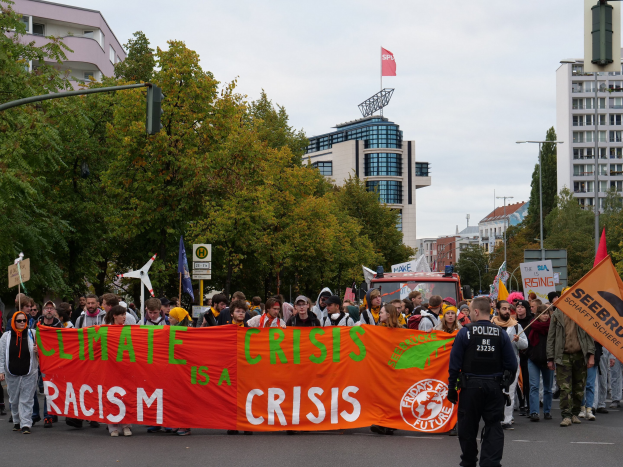 Menschen marschieren mit einem "Klima-Krise ist eine Krise"-Schild die Straße entlang, mit Bäumen, Straßenschildern, einem geparkten Fahrzeug, Gebäuden und einem blauen Himmel im Hintergrund.