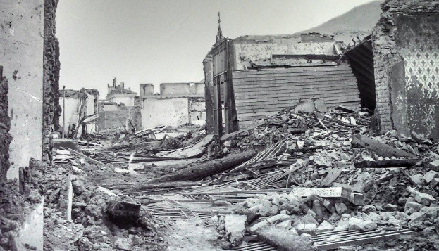 Ein Schwarz-Weiß-Foto eines schwer beschädigten Gebäudes auf einer Straße, umgeben von Trümmern wie eingestürzten Wänden, Holzstöcken und Steinen, mit einem Hügel und Himmel im Hintergrund.