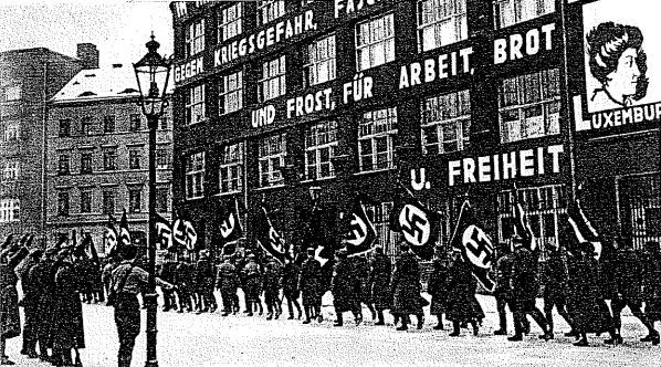 Black and white image of a group marching down a street holding flags and banners, with a light pole on the left and a board reading "German Nazi Party in Berlin" on the right.