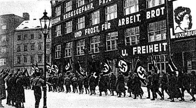 Black and white image of a group marching down a street holding flags and banners, with a light pole on the left and a board reading "German Nazi Party in Berlin" on the right.
