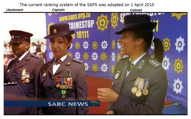 Eine Gruppe von Menschen in Uniformen und Hüten steht vor einem Banner mit der Aufschrift "Das aktuelle Rangsystem der SAP wurde am 1. April 2010 übernommen" oben und unten auf dem Bild.