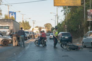 Eine Gruppe von Menschen steht um ein verunglücktes Motorrad auf der Straße mit mehreren Fahrzeugen, darunter ein Lkw, und einem Hintergrund aus Bäumen, Polen, Lampen und Schildern unter dem Himmel.