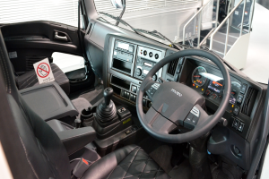 Ein Truck-Interieur mit Lenkrad, Armaturenbrett, Sitzen, Ganghebel, Tacho, Scheibenwischer, Seitenspiegel und weiteren Objekten, mit Geländern und anderen Fahrzeugen im Hintergrund.