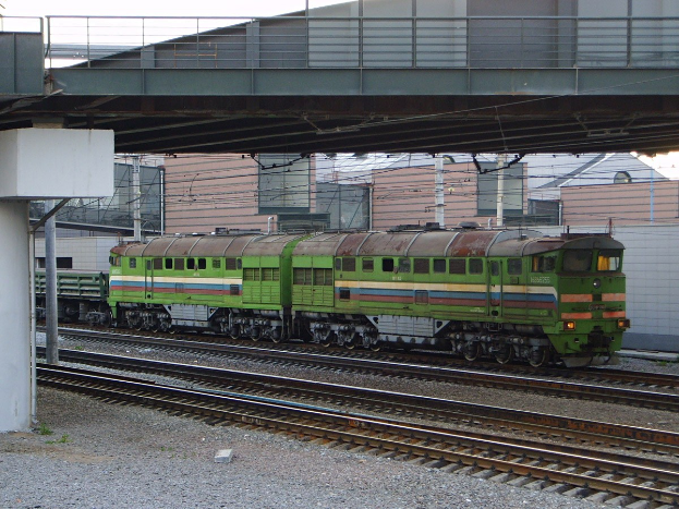 Grüne Diesel-Elektrolokomotive auf Schienen unter einer Brücke, mit einem Steinpfeiler links, Gebäuden und Strommasten im Hintergrund und einem klaren blauen Himmel.