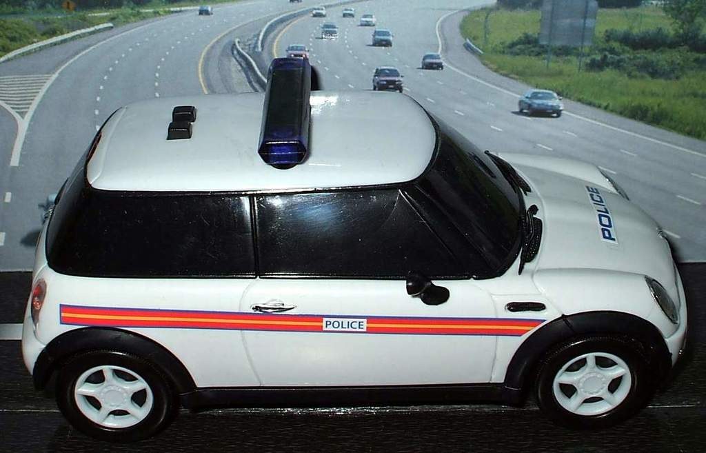 White police car with "Police" markings in front of moving traffic on a road lined with grass and trees outside a city.