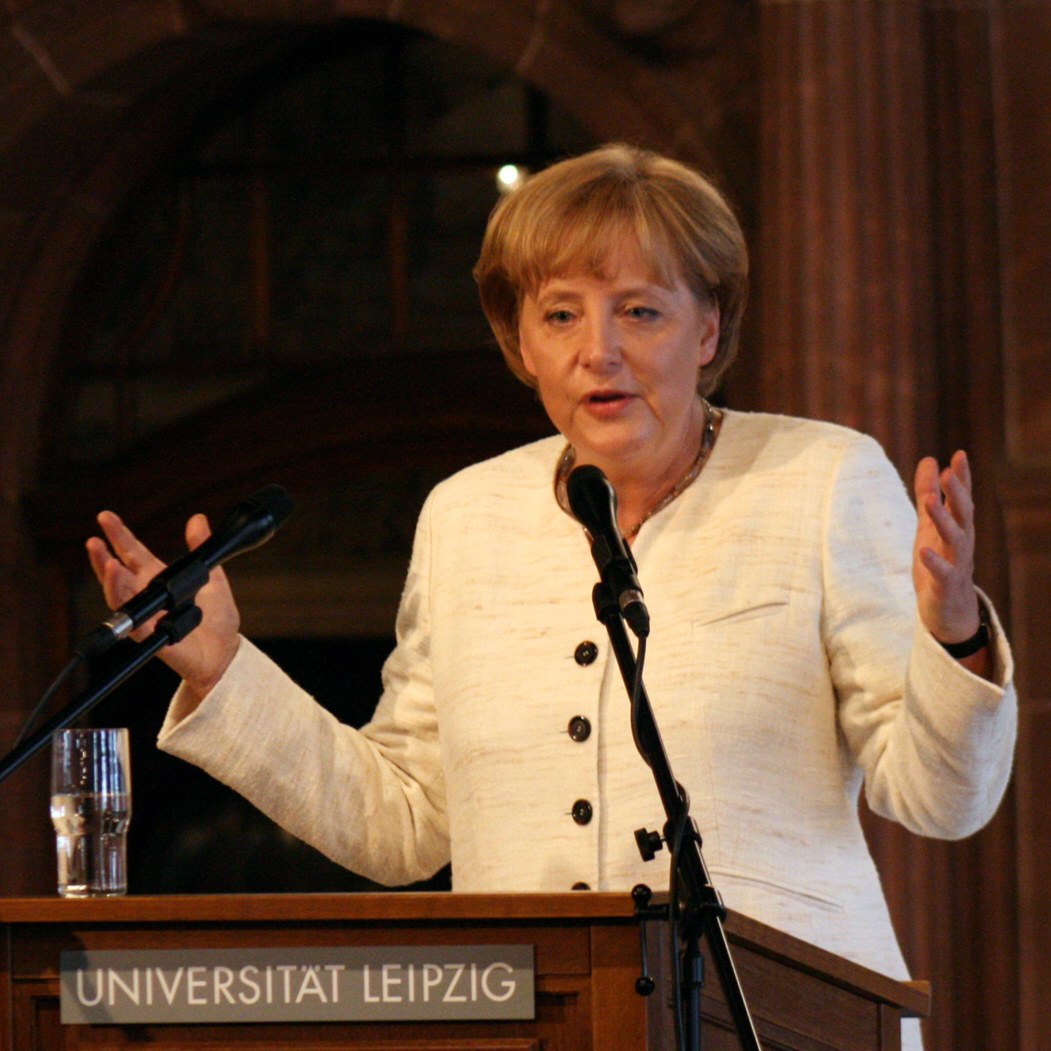 German Chancellor Angela Merkel speaking at a podium with a microphone and a glass of water at the University of Leipzig.