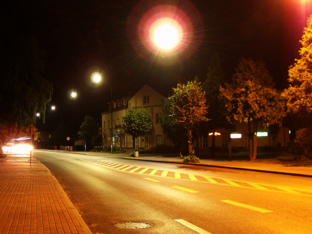 Eine nächtliche Straßenlaterne in einer Stadt mit beleuchteter Straße, einem Gehweg auf der linken Seite, Bäumen und Gebäuden im Hintergrund und einem dunklen Himmel darüber.