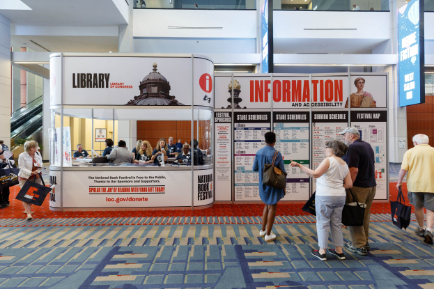 Eine Gruppe von Menschen versammelt sich vor einem Stand mit Schildern, einer Anzeigetafel und Sitzgelegenheiten, einige halten Taschen.