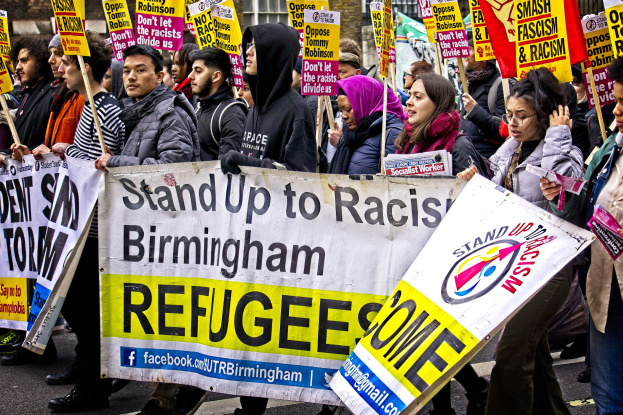 Group of people marching in a protest or demonstration, holding signs and banners, with a building in the background.