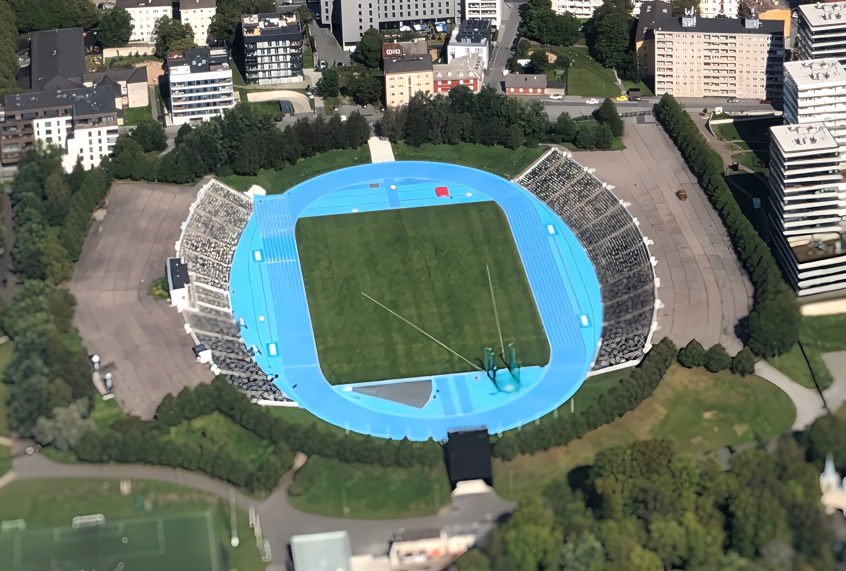 Aerial view of the Olympic Stadium in Munich, Germany, surrounded by city buildings, trees, roads, vehicles, and grass.