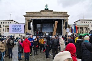 Eine Menschenmenge steht vor einem Gebäude mit einer Bühne, Lautsprechern und einem Bildschirm, hält Fahnen und Transparente, was auf eine Demonstration in Berlin hindeutet.