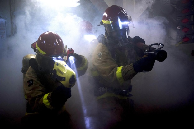 Zwei Feuerwehrleute in Schutzausrüstung sprühen Wasser auf eine rauchgefüllte Wand, mit einer Tafel und Gegenständen in der Nähe.