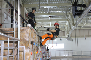 Zwei Personen in schwarzen und orangen Outfits und Helmen klettern an einer Felswand in einer Halle, gesichert mit Seilen, mit einem Metallgeländer links und einer weißen Wand mit Lampen im Hintergrund.