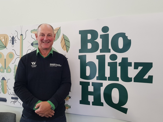 Ein lachender Mann, identifiziert als Richard Geddes, CEO von Bio Blitz HQ, steht vor einem "Bio Blitz HQ"-Schild mit einem Tisch und einem Stuhl links daneben und einer Tafel mit Text und Bildern an der Wand dahinter.