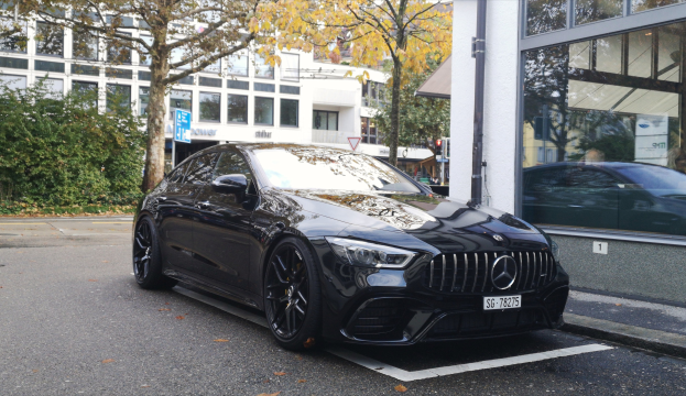 Schwarzer Mercedes-AMG GT 63 S 4MATIC+ auf einer Straße mit Glasfenstergebäuden, Bäumen und städtischer Infrastruktur im Hintergrund.