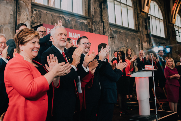 A group of people clapping in celebration in front of a crowd, with a podium, microphone, and board with text on the right, and chairs, a banner, wall, windows, and lights in the background.