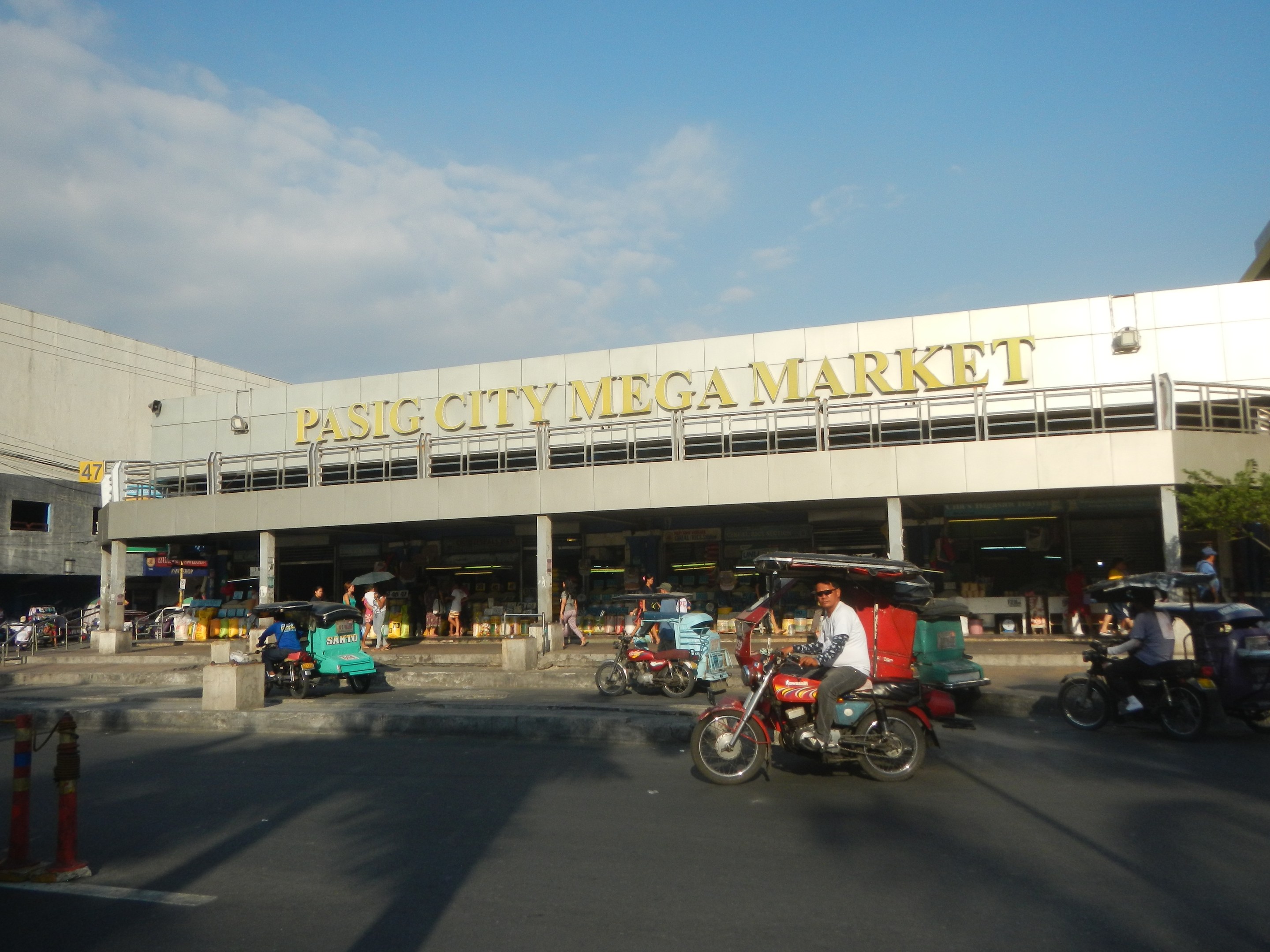 Lebendiger Pasig City Mega Markt mit Fahrzeugen auf der Straße, Fußgängern, einem Gebäude mit Text im Hintergrund und einem bewölkten Himmel.