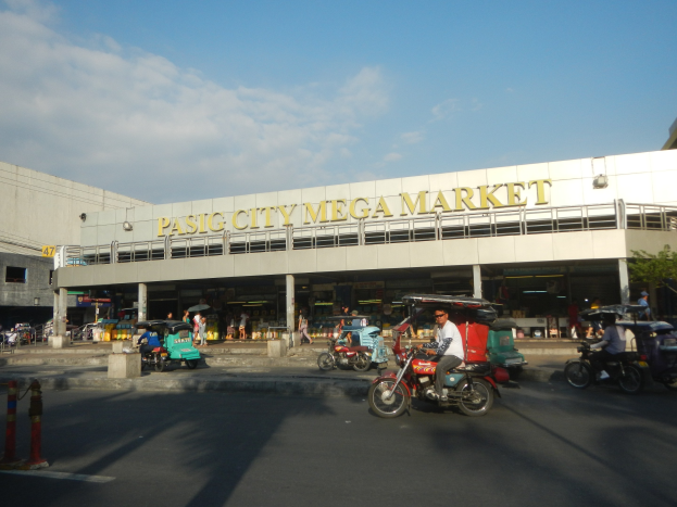 Lebendiger Pasig City Mega Markt mit Fahrzeugen auf der Straße, Fußgängern, einem Gebäude mit Text im Hintergrund und einem bewölkten Himmel.
