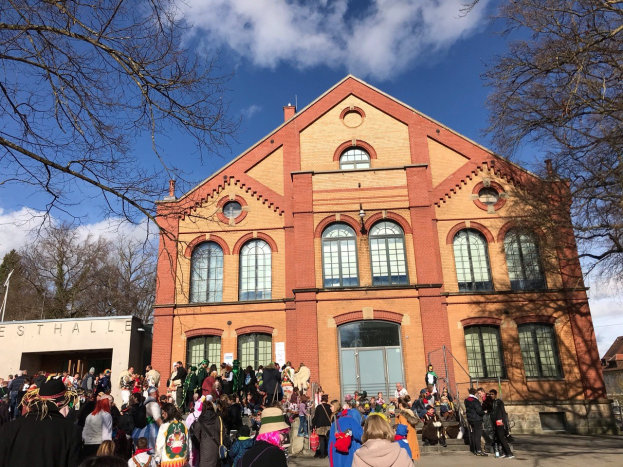 Eine große Gruppe von Menschen steht vor einem mehrstöckigen Gebäude mit Fenstern und Türen, umgeben von Bäumen und einem bewölkten Himmel, einige tragen Mützen und tragen Taschen.