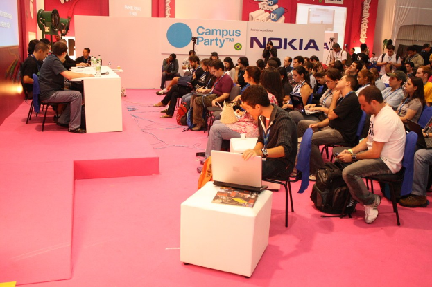 Conference with seated attendees listening to speakers on stage, media and owners conversing, with a large hoarding and wall in the background.
