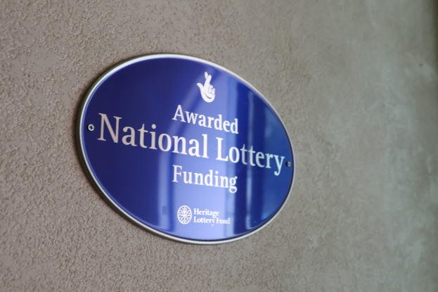Blauer rechteckiger Schild an einem Gebäude mit weißem Hintergrund und fetter schwarzer Schrift, die "National Lottery Funding awarded" lautet.