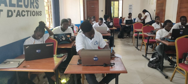 Eine Gruppe von Schülern in einem Klassenzimmer mit Laptops, Taschen auf dem Boden und einer Wand mit Text, die einen Online-Kurs in Ghana anzeigt.