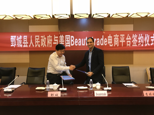 Zwei Männer, die vor einem Tisch mit Namensschildern, Mikrofonen, Flaschen, Tassen, Papieren und anderen Gegenständen stehen und ein Memorandum of Understanding (MOU) zwischen Beautetrade und China unterzeichnen, mit einem Banner und Lichtern im Hintergrund.