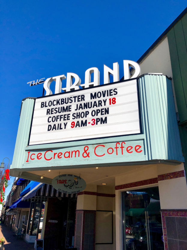 The Strand Blockbuster Movies Resume January 18 Coffee Shop building with a name board, a street pole, a group of trees, some plants in pots, and a cloudy sky.