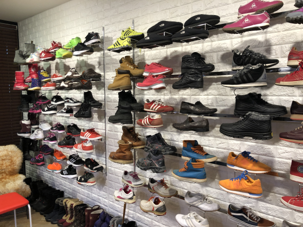 Ein Schuhgeschäftsinneres mit zahlreichen Schuhen auf Gestellen, einem Stuhl mit einem Teddybär auf der linken Seite und einer Wand im Hintergrund.