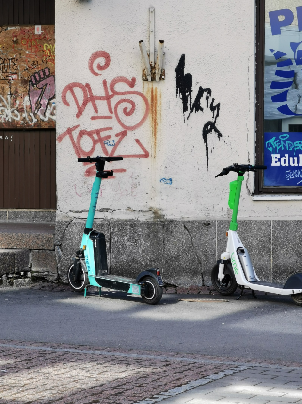 Zwei Elektro-Scooter nebeneinander auf einer Straße mit einer Graffiti-bedeckten Wand und einem Plakat im Hintergrund.