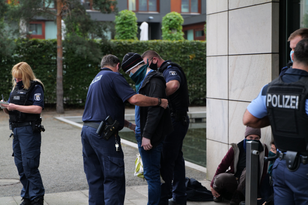 Gruppe von Polizeibeamten vor einem Gebäude stehend, mit einer Frau, die ein Buch und einen Stift hält, und mehreren Personen in der Nähe sitzend und stehend, vor dem Hintergrund von Pflanzen, Bäumen und Gebäuden.