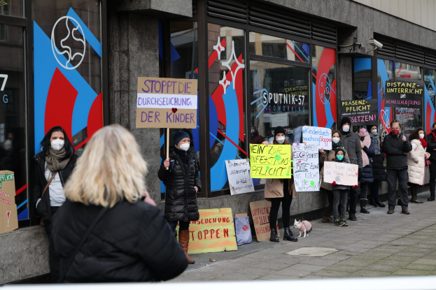 Eine Gruppe maskierter Menschen protestiert vor einem Glasfrontgebäude und hält Schilder und Plakate hoch, während Kameras und ein Hund anwesend sind.