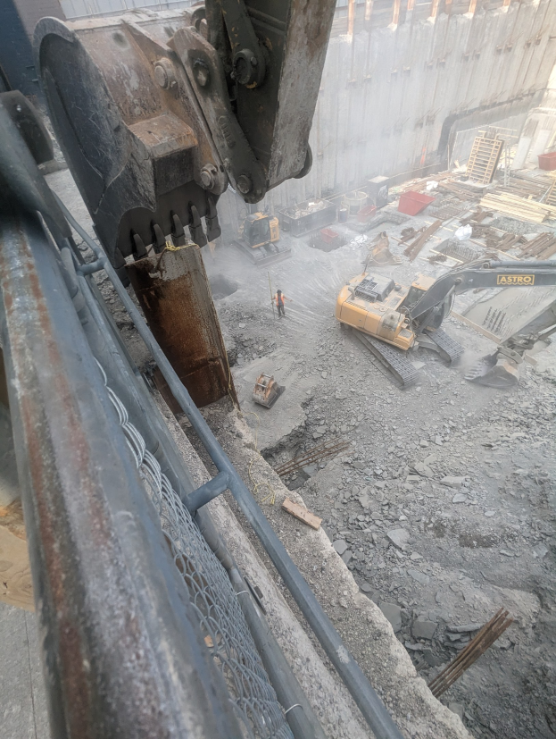 Baustelle mit großem Bagger beim Abriss eines Gebäudes, mehrere Fahrzeuge und Personen sowie ein Metallobjekt links.