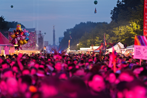 Eine große Menschenmenge auf einem nächtlichen Musikfestival, beleuchtet von farbigen Lichtern und Ballons, mit Zelten, Bannern, Fahnen, Bäumen, einem Turm und einem sternenklaren Himmel im Hintergrund.