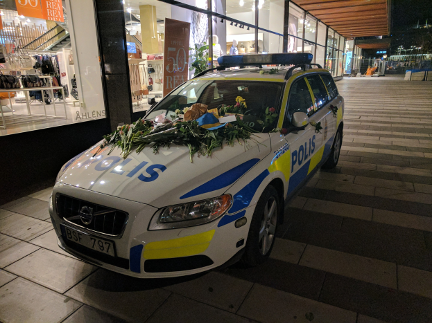 Ein Polizeiwagen mit Blumenverzierung parkt nachts vor einem Geschäft mit Glaswänden, auf denen Schaufensterpuppen in Kleidern und beleuchtete Schilder zu sehen sind.