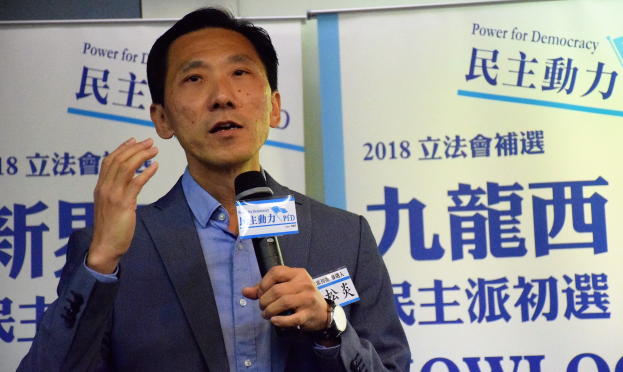 A suited man speaking into a microphone at an event with banners in the background.