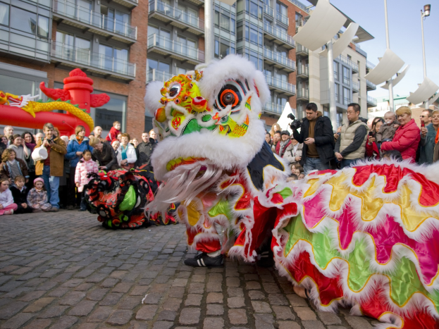 Ein lebendiges chinesisches Neujahrsfest in Amsterdam mit einer Löwen-Tanzvorstellung und einer Zuschauermenge, einige machen Fotos, vor einem Hintergrund aus Gebäuden, Laternenmasten und einem klaren blauen Himmel.