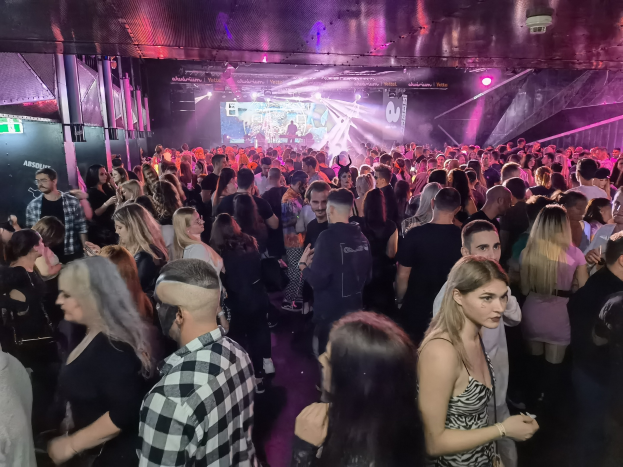 Eine große Menschenmenge in einer Diskothek mit Deckenbeleuchtung und Säulen, mit einem Bildschirm im Hintergrund, der ein Absolut Nightlife-Event anzeigt.