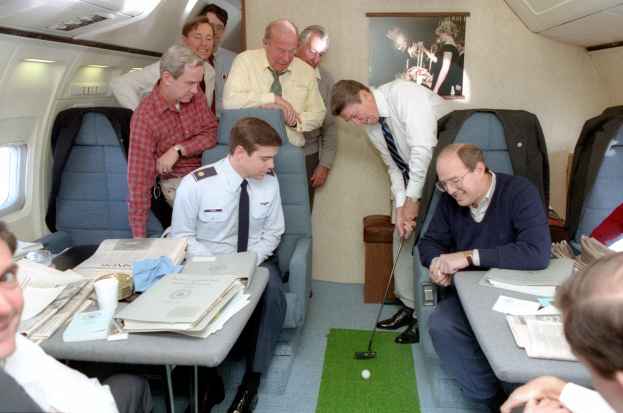 A group of men sitting around a table in an airplane, with books, papers, cups, and other objects on the table, a photo frame on the wall, ceiling lights, and one man holding a golf club.