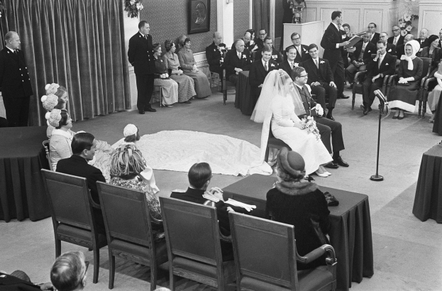 Schwarzes und weißes Foto einer Hochzeitszeremonie mit Menschen, die sitzen und stehen, einem Mann mit Mikrofon, einer Frau mit Blumenstrauß, einem Podium mit Mikrofon, Blumenschmuck, einem Vorhang, einem gerahmten Foto an der Wand und einer Tür im Hintergrund.