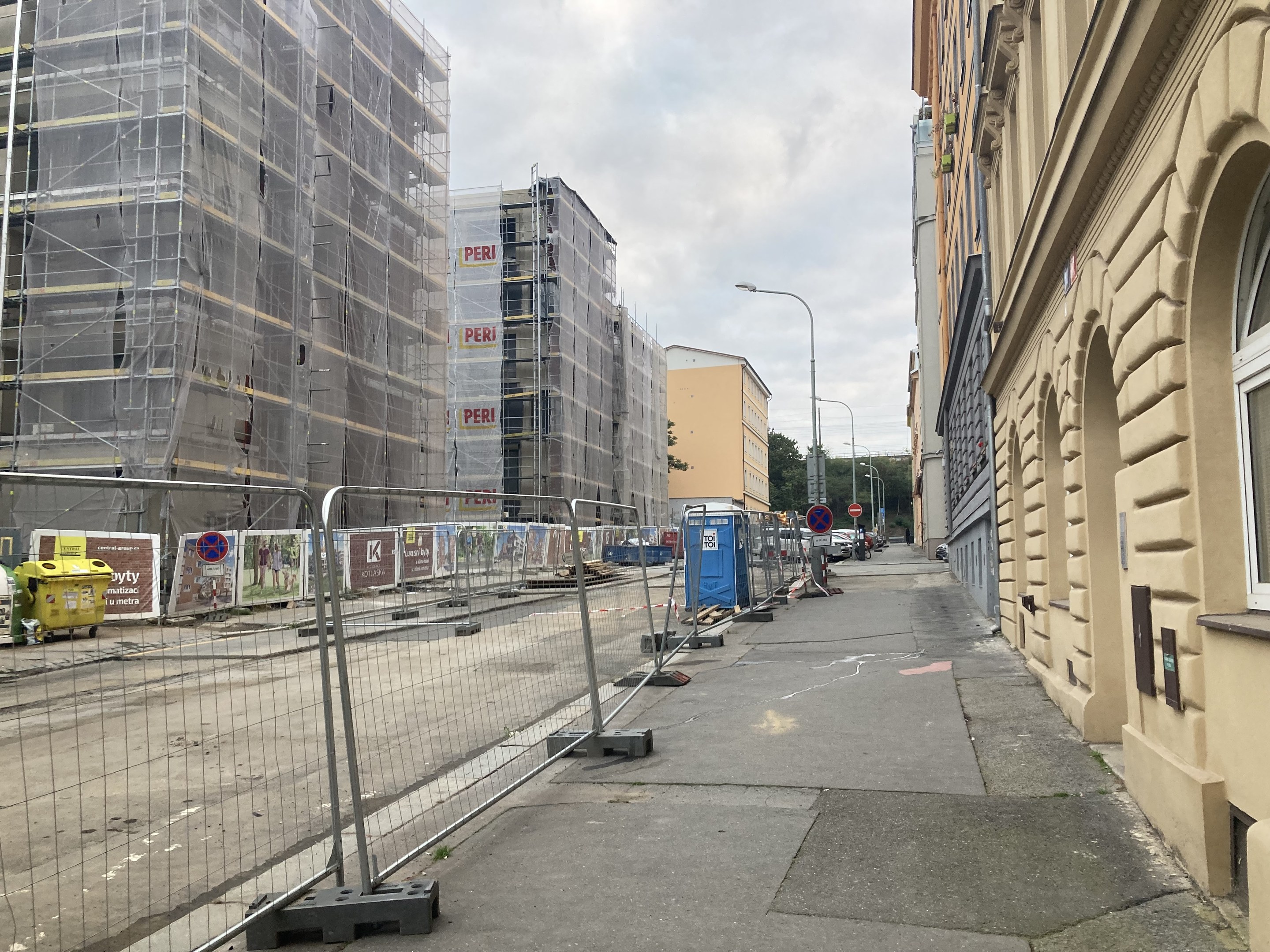 A city street with buildings, street poles, lights, vehicles, barrier poles, signboards, bins, trees, and a cloudy sky, featuring a construction site with scaffolding in the foreground.