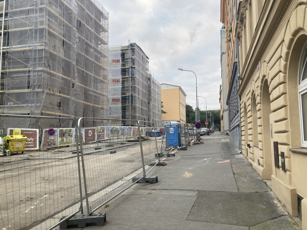 A city street with buildings, street poles, lights, vehicles, barrier poles, signboards, bins, trees, and a cloudy sky, featuring a construction site with scaffolding in the foreground.