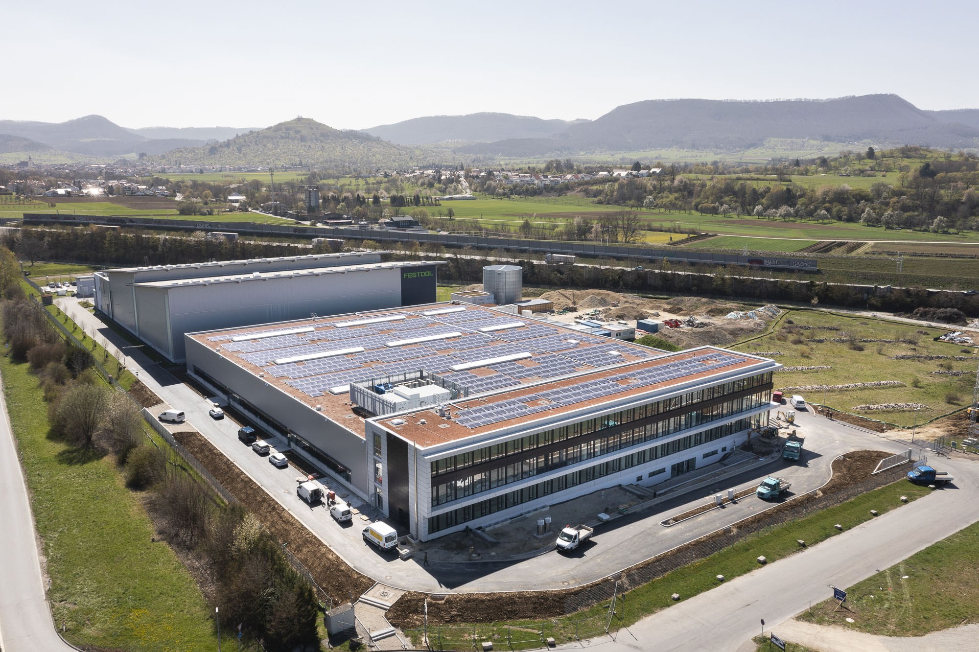 Aerial view of a large building with solar panels on its roof, surrounded by vehicles, trees, grass, and poles, with hills and a clear blue sky in the background; the building is Germany's first solar-powered factory.